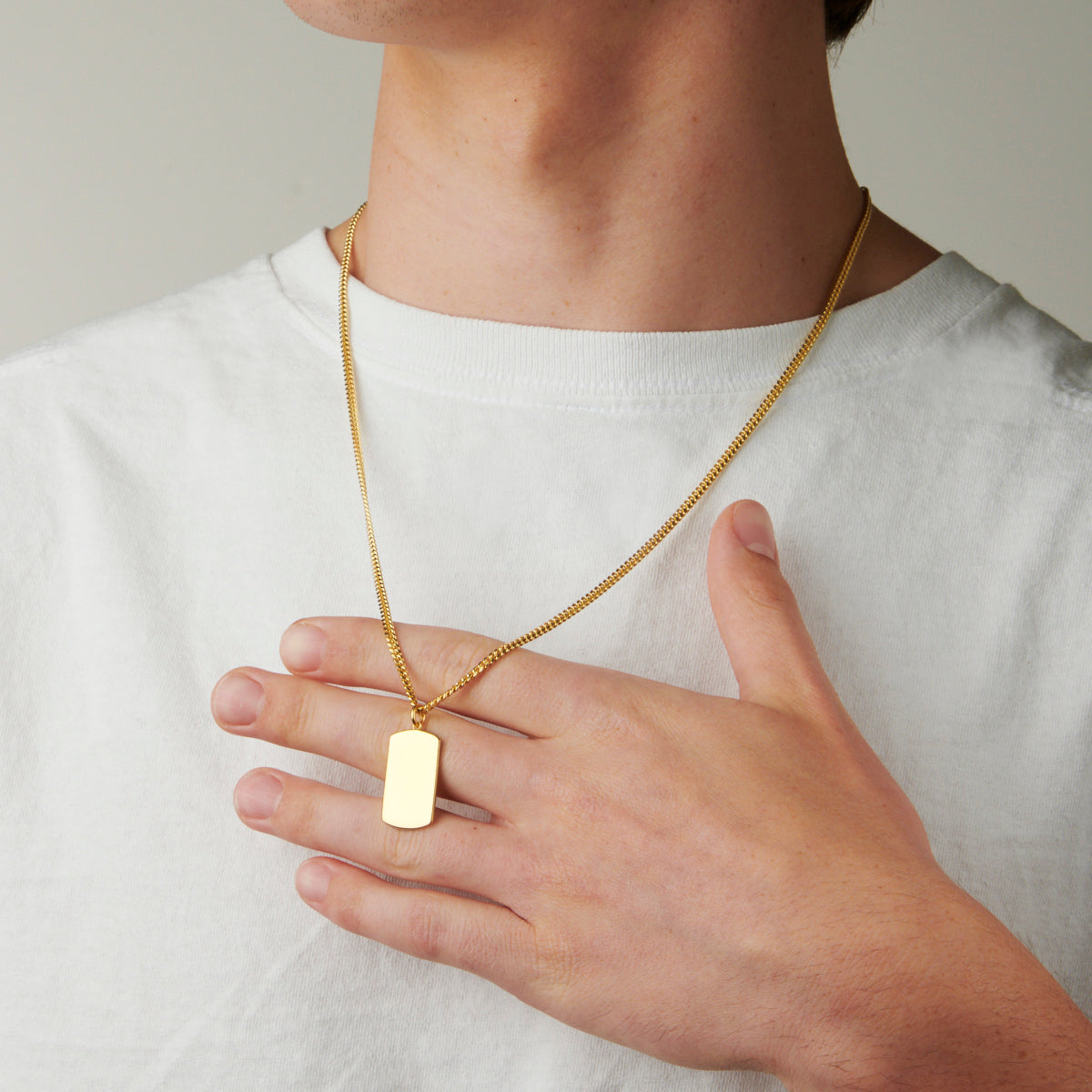 A person wearing the Classic Large Tag Pendant, with the pendant hanging on a 20" or 22" tub chain around their neck. The pendant is available in sterling silver, gold vermeil, or 14kt yellow gold and can be engraved with custom font and text. The person in the image is dressed in casual attire and the pendant adds a touch of elegance and personalization to their outfit.